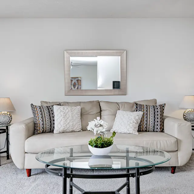 A light-colored living room featuring a beige sofa with decorative pillows, two stylish side tables with lamps, a glass coffee table, and a large mirror on the wall.