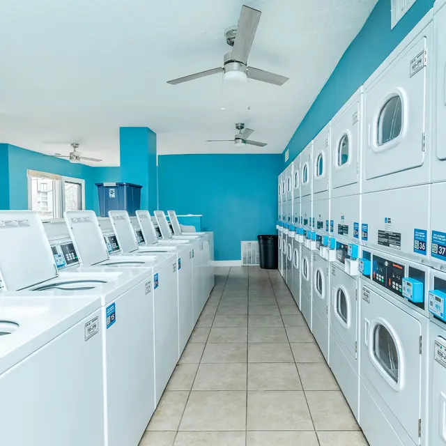 A modern laundry room with several white washing machines and dryers aligned along the walls. The walls are painted a bright blue and the floor is tiled. Ceiling fans are visible.