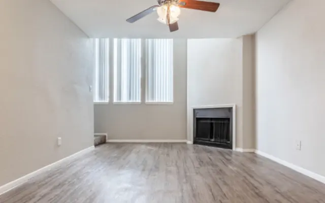 Empty Living Room With Fireplace A spacious, empty living room featuring a ceiling fan, light wood flooring, and a fireplace. Large windows allow natural light to fill the room.