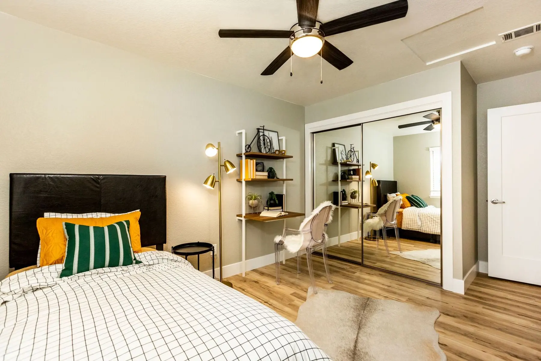 A modern bedroom featuring a black bed frame, patterned bedding, a ceiling fan, and a mirrored closet. There is a small desk with decorative items against the wall and a door leading to another room.