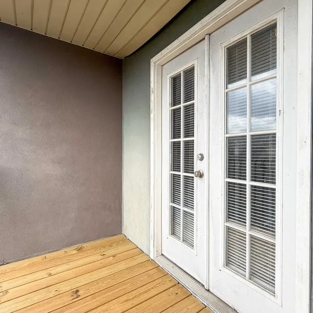A small balcony with a wooden floor and double French doors leading inside. One side of the balcony has a gray wall, while the other side is painted a light color. The sky is visible through the doors, hinting at an overcast day.