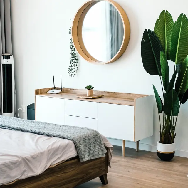 A modern bedroom featuring a wooden bed, circular mirror, sleek dresser, and a tall potted plant against a light wall.