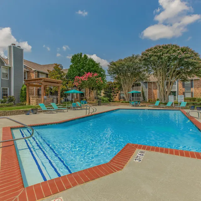 A sunny outdoor swimming pool in an apartment complex, surrounded by green trees and lounge chairs.