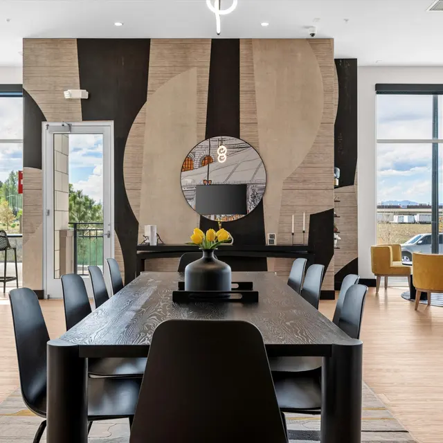 A modern dining area featuring a long black table set with a centerpiece, surrounded by dark chairs, an artistic wall in the background, and large windows with a view of the outdoors.