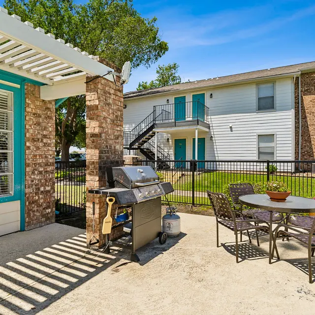 An outdoor patio area featuring a charcoal grill and a dining table with chairs, situated next to a brick structure and an apartment building in the background.