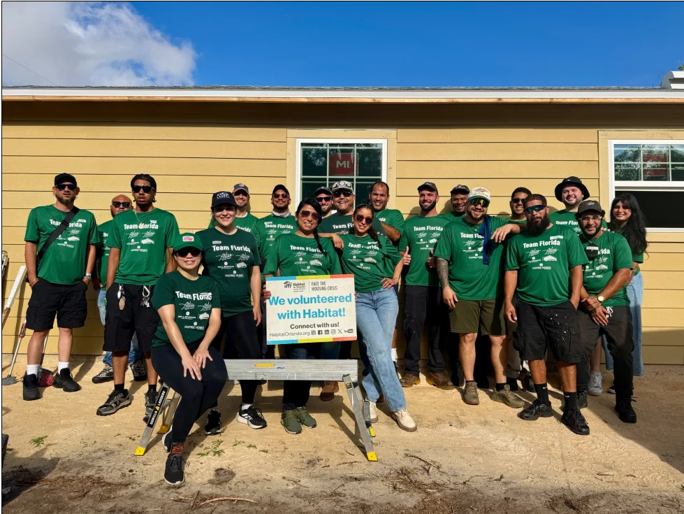 Group of volunteers wearing matching green shirts in front of a house, holding a sign that says 'We volunteered with Habitat!'