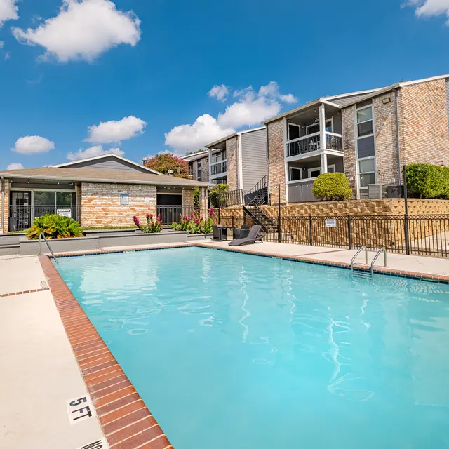A clear swimming pool surrounded by a securely fenced area in an apartment complex. The pool area has lounge chairs and is framed by modern buildings under a blue sky with fluffy clouds.