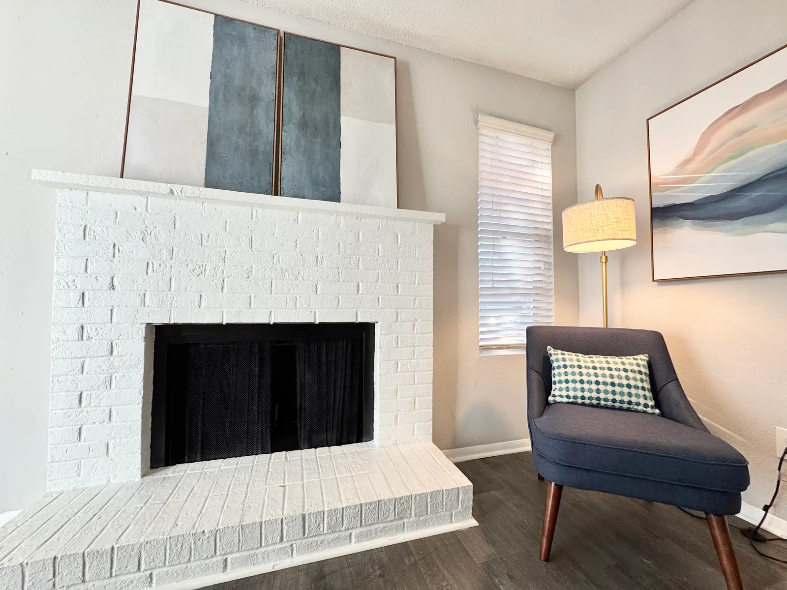 Cozy living room featuring a white brick fireplace, modern artwork, and a blue accent chair with a patterned cushion.
