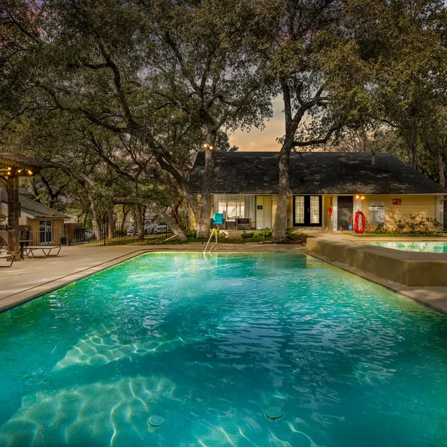 A serene swimming pool area with clear water, surrounded by trees and lounge chairs under a twilight sky. A building is visible in the background, with warm ambient lighting.