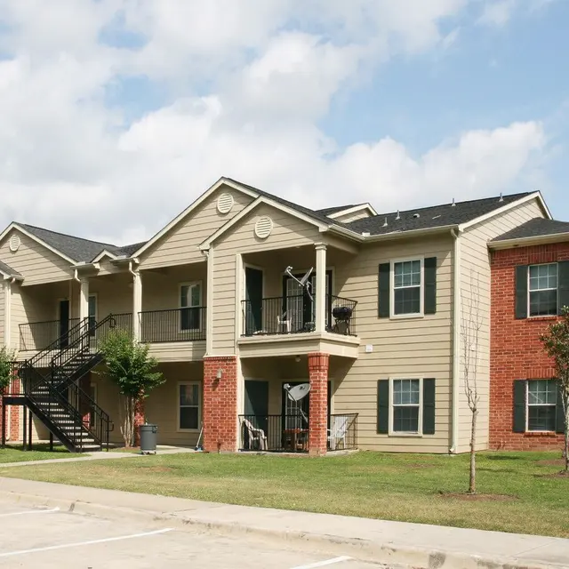 Apartment Complex Exterior A two-story apartment complex with a combination of beige and red brick exteriors, featuring balconies and stairs, surrounded by green lawn and trees.