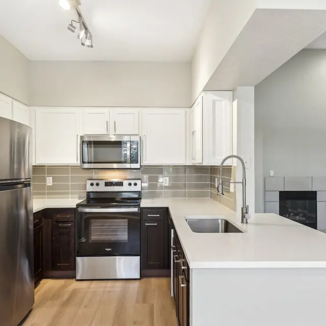 Stylish Modern Kitchen A modern kitchen featuring stainless steel appliances, including a refrigerator, stove, and microwave, with dark wood and white cabinetry. The countertop is light-colored and there is a sink with a modern faucet. A living area with a fireplace is visible in the background.
