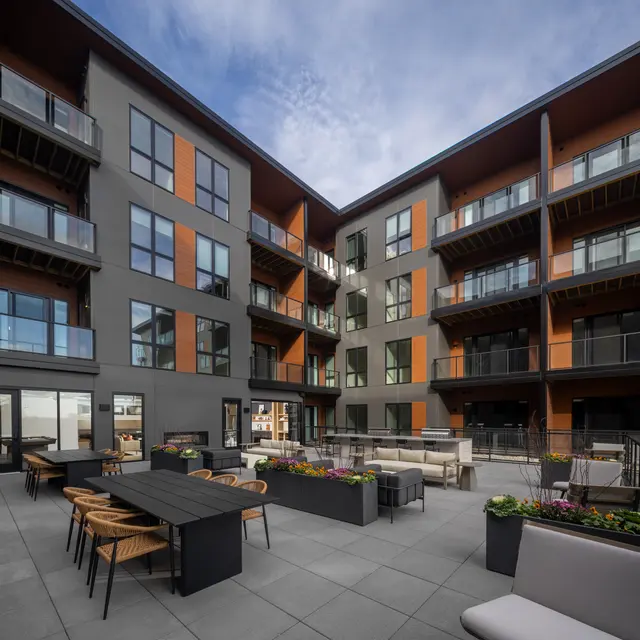 A modern apartment courtyard featuring gray and orange facades, balconies, and outdoor furniture arranged around planters filled with flowers.