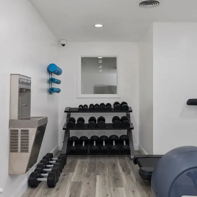 A well-lit gym space featuring free weights, a treadmill, and a water fountain. The walls are painted white, and the flooring is wood-like with a clean, modern aesthetic.