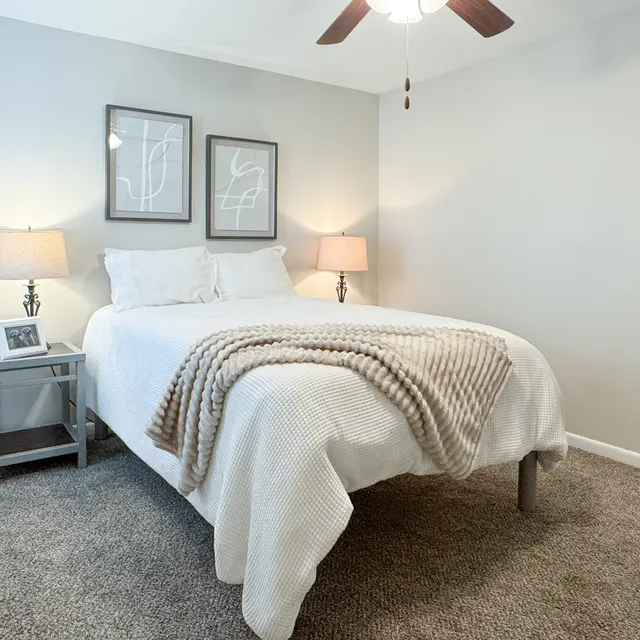 A cozy bedroom featuring a queen-sized bed with a beige blanket, two bedside lamps, and framed artwork on the wall. The room has light gray walls and a carpeted floor.