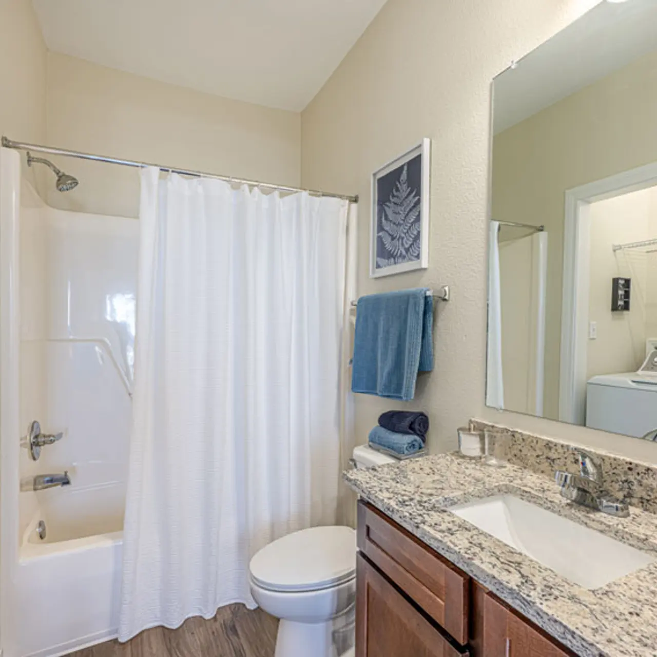 A modern bathroom featuring a shower tub with a white curtain, a toilet, and a sink with granite countertops. A laundry area is visible in the background.