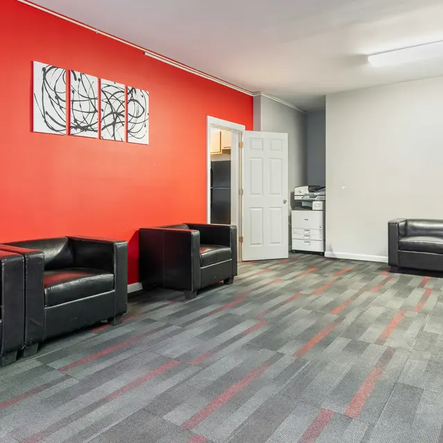 A modern office lounge area featuring black leather chairs, a vibrant red accent wall, and a gray wall. The flooring is patterned in gray and colorful tiles. A door leads to another room and a printer is visible in the background.