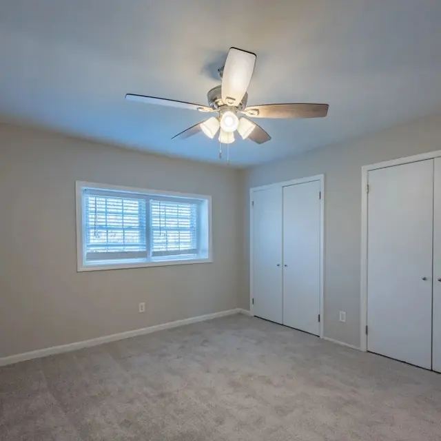 A clean, empty bedroom featuring a ceiling fan, carpeted floor, and a window with blinds. Two closet doors are visible on the right side of the image.