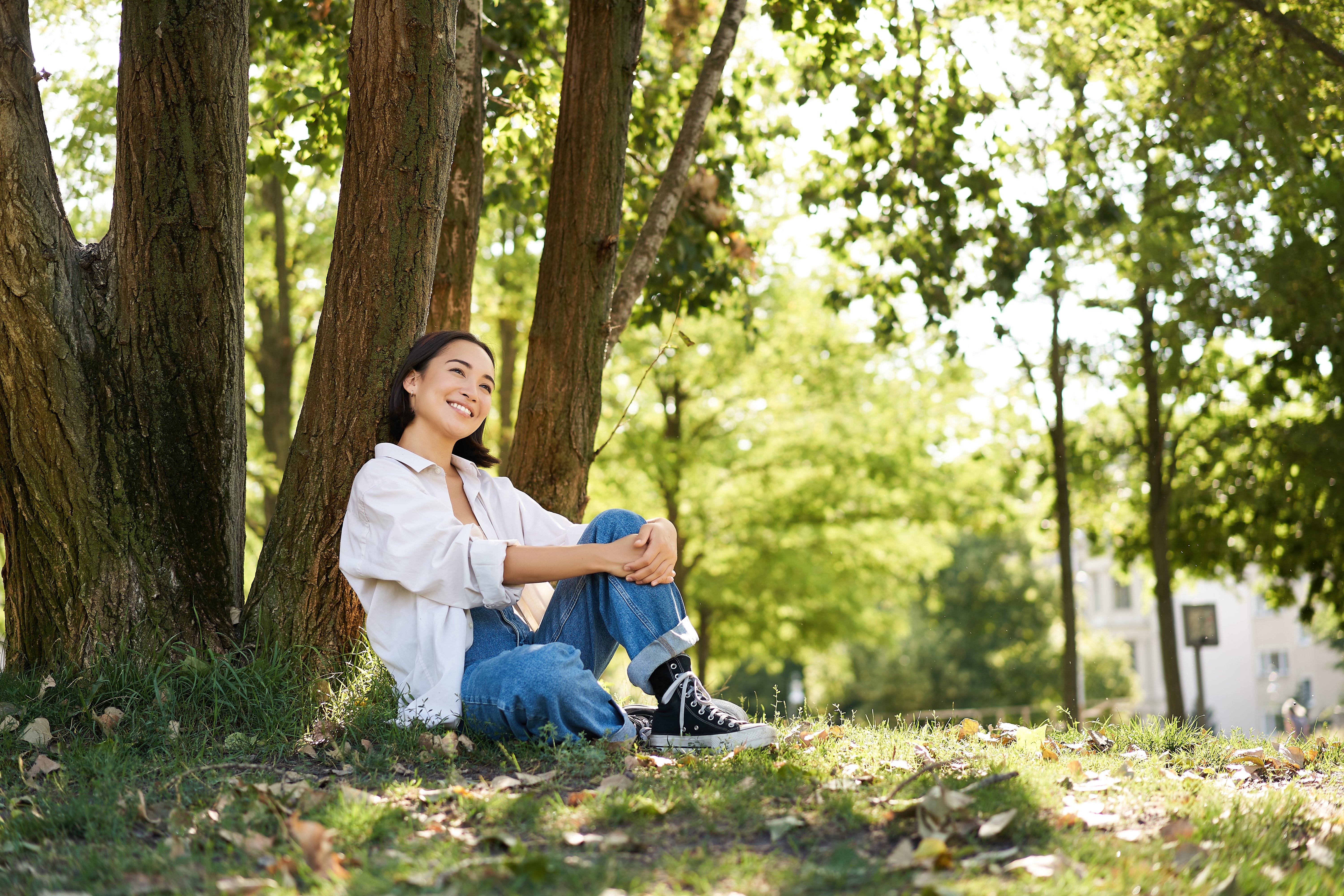 A young woman sits on the grass beneath a tree, smiling and looking relaxed. She is dressed casually in a white shirt and jeans, with her hair tied back.