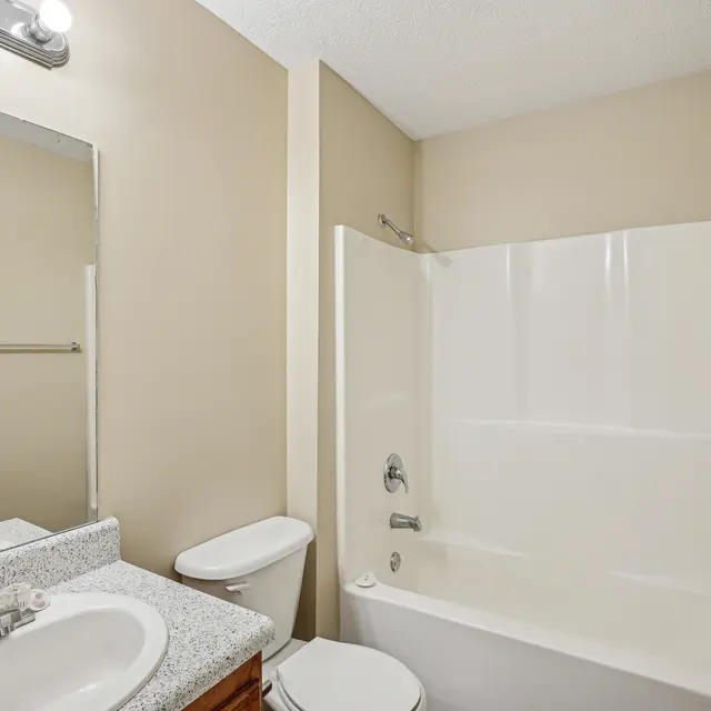 A clean, modern bathroom featuring a tub with shower, a toilet, and a sink with a granite countertop. Beige walls and lighting enhance the bright atmosphere.