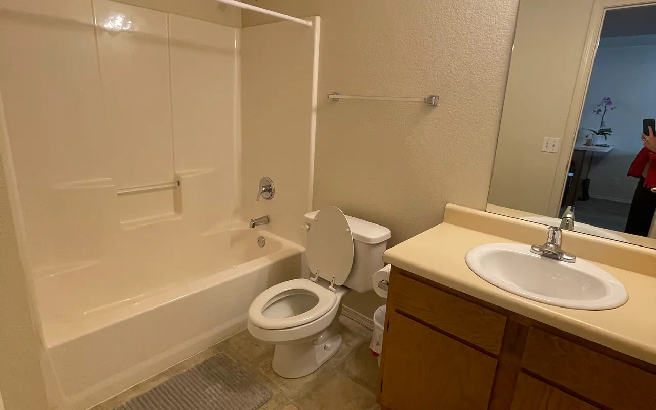 A clean and well-lit bathroom featuring a bathtub with a shower, a toilet, and a vanity with a sink. The floor is tiled, and bath mats are placed in front of the bathtub and toilet. A large mirror is mounted above the sink with wall sconces for lighting.