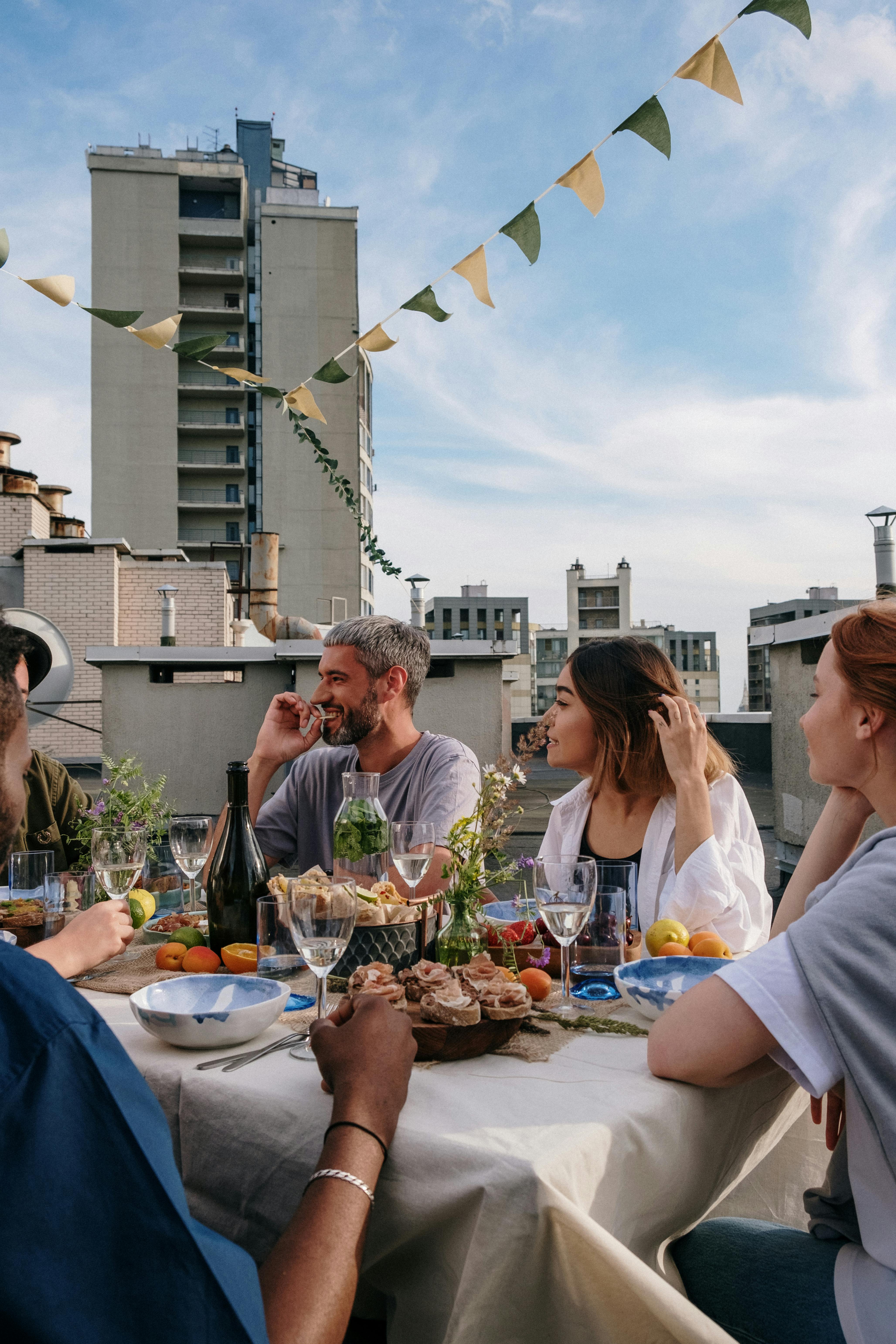 Rooftop Dinner Gathering A group of people enjoying a meal at a rooftop setting, surrounded by urban buildings and decorative bunting. The table is set with various dishes, drinks, and fresh fruits, creating a vibrant atmosphere.