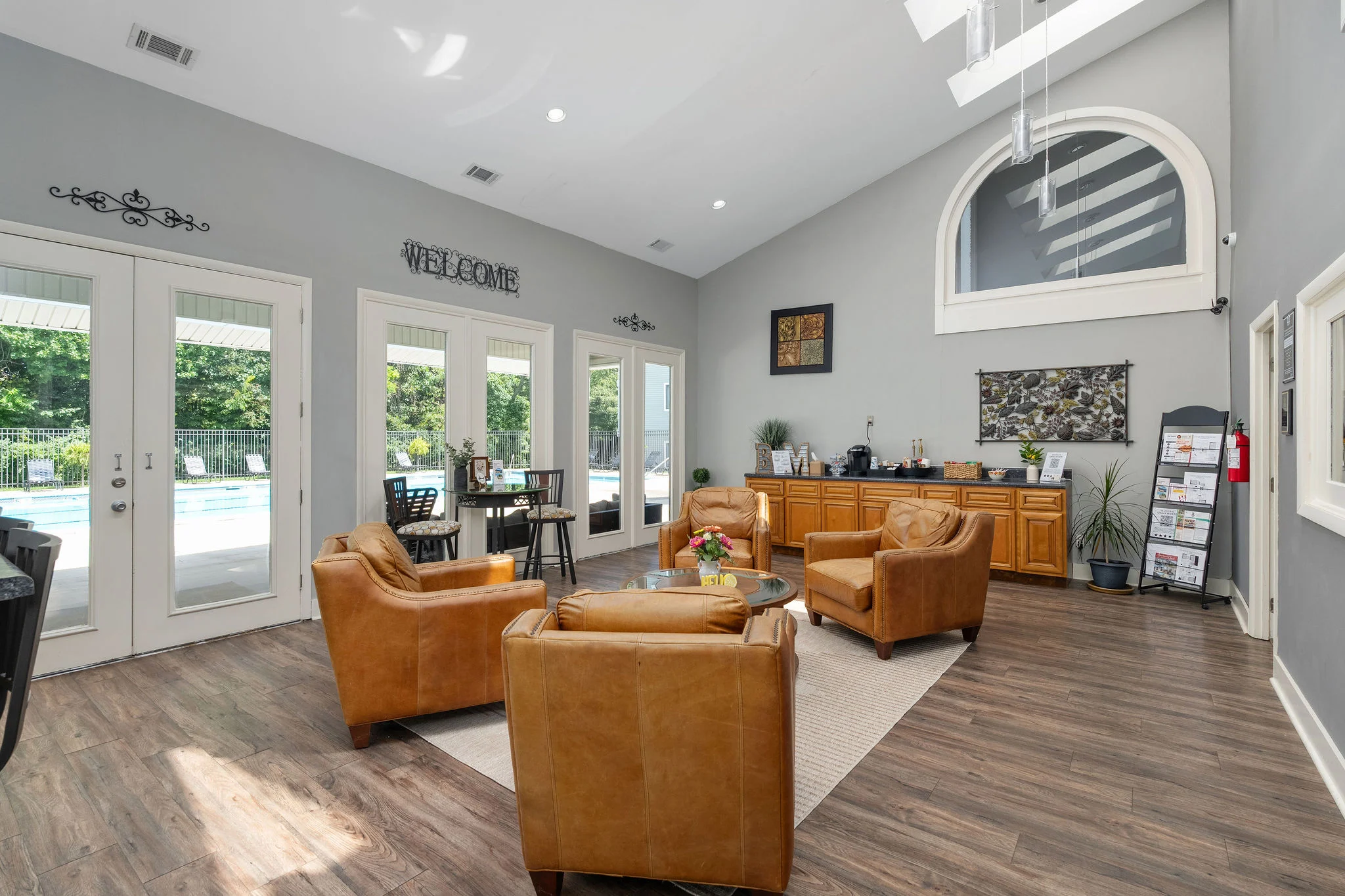 A welcoming community lounge area with brown leather chairs, a coffee table, and a kitchenette in the background. Large windows let in natural light, and the space has a relaxed atmosphere.