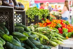 Fresh Vegetables at Farmers Market A vibrant display of fresh vegetables at a farmers market, including cucumbers, corn, and bell peppers, with various customers in the background.