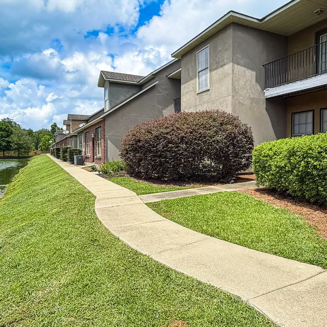 Pathway next to a pond with apartment buildings on the right, surrounded by greenery and blue sky with clouds.