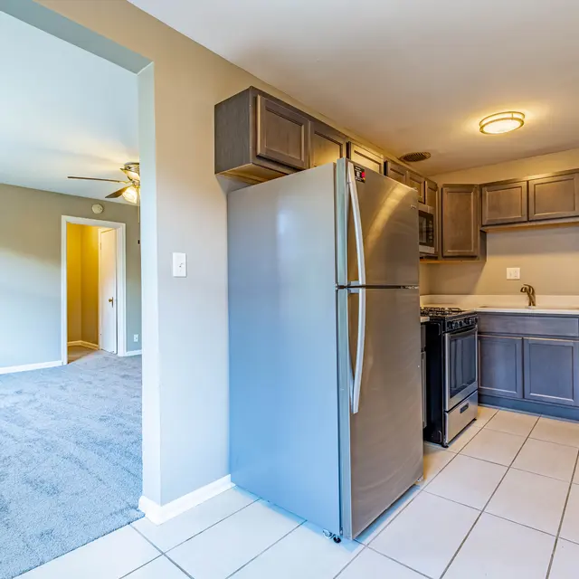 A kitchen area with dark cabinetry and stainless steel appliances, including a refrigerator and stove, facing an adjacent living room with gray carpet. The walls are painted beige, and there is a doorway leading to the living room.