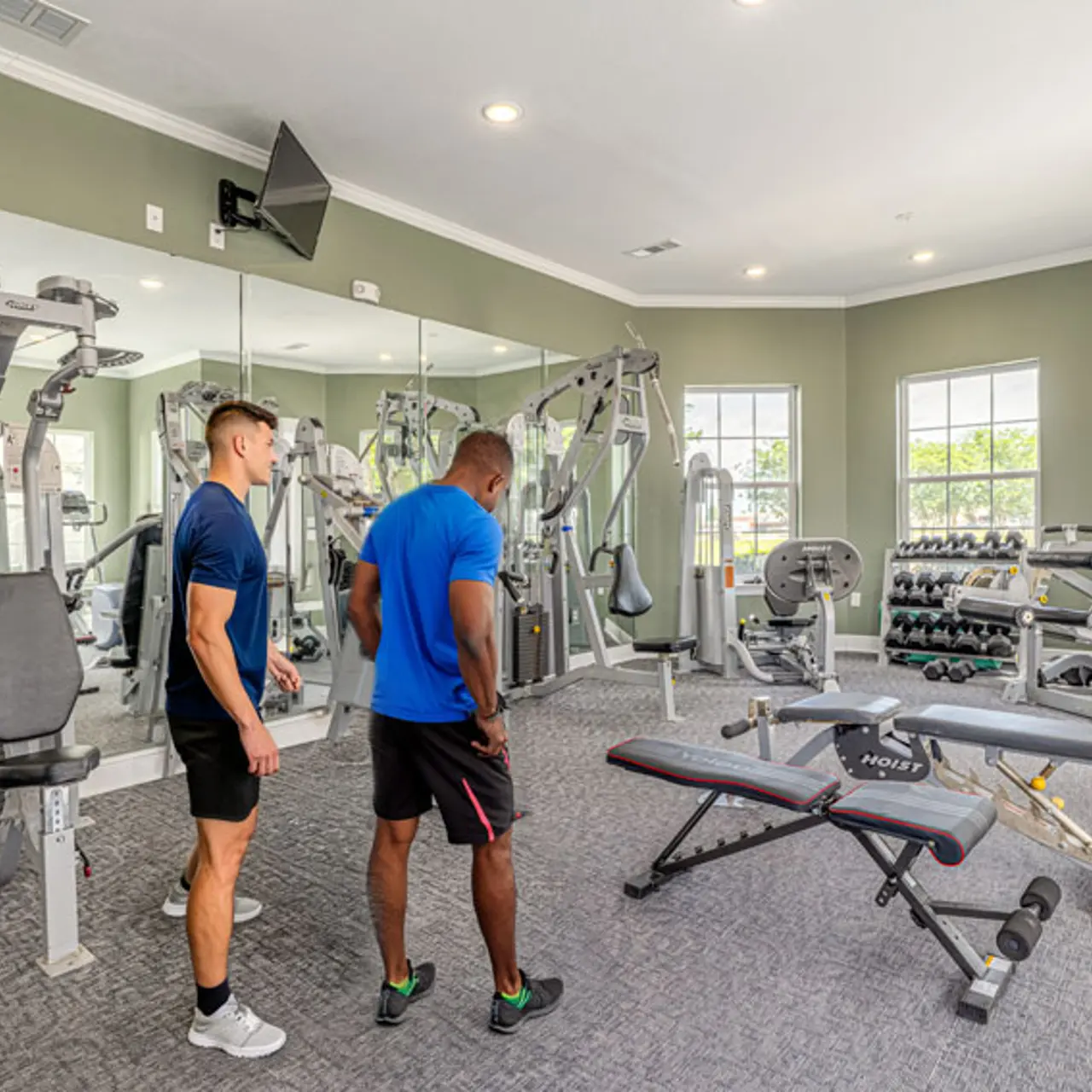 Two men standing in a modern gym with various exercise equipment. One man is wearing a blue shirt and the other a black shirt, observing a weight bench in front of them.
