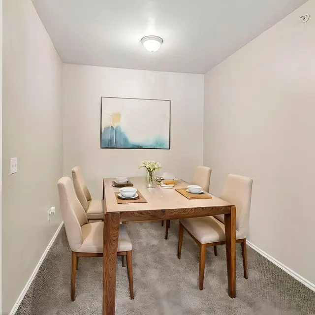 Cozy Dining Space A dining area featuring a wooden table with four chairs, set for a meal with plates and a vase of flowers, against a light-colored wall with an abstract painting.