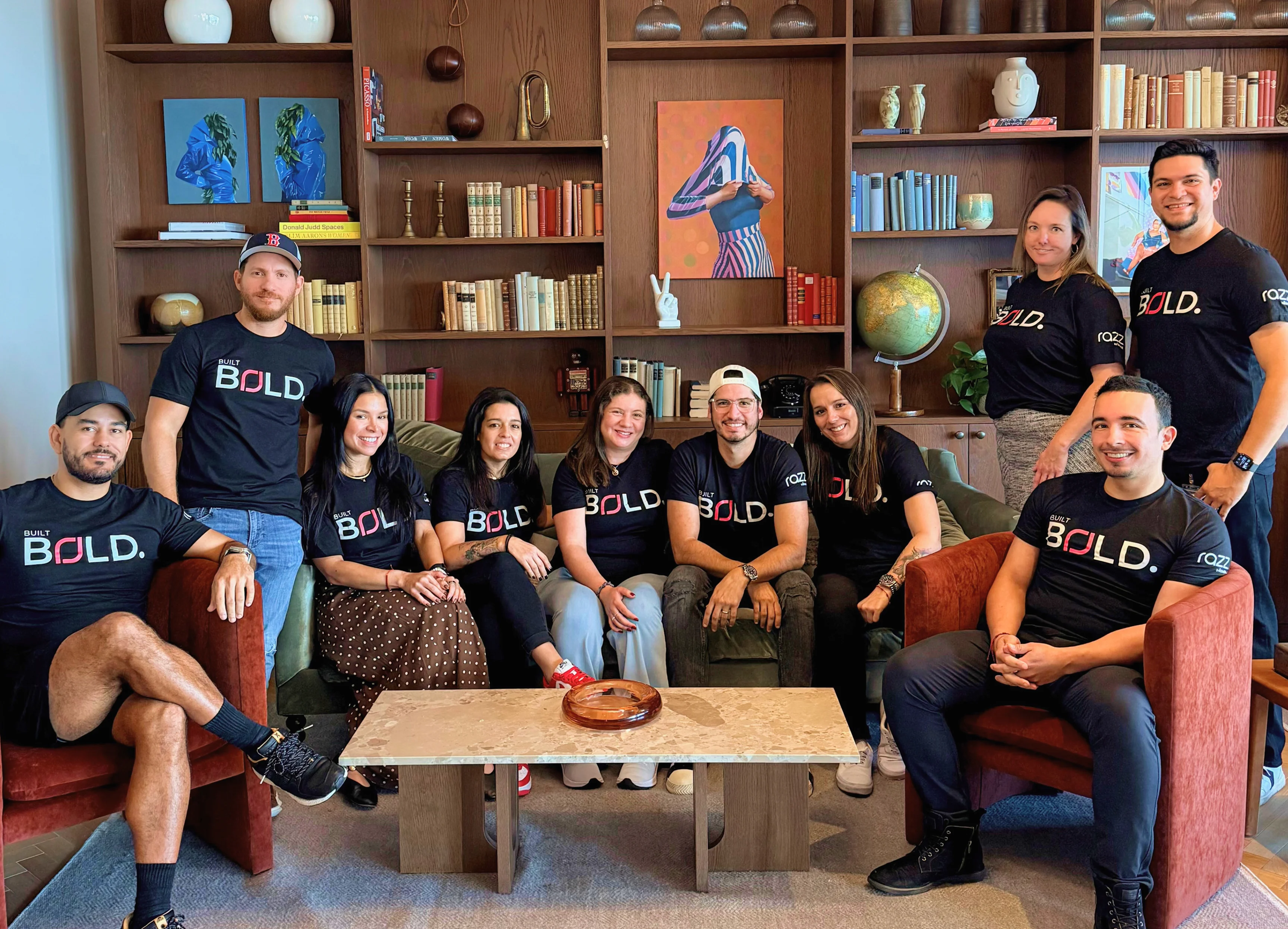 BOLD Team Group Photo A group of eleven individuals sitting and standing around a coffee table in a stylish indoor setting, wearing matching black t-shirts with the word 'BOLD' printed on them. They are arranged in two rows, some seated on chairs and others on a sofa, with bookshelves and decorative items in the background.