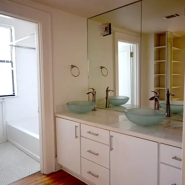 A modern bathroom featuring a double sink vanity with glass bowls, a mirror extending across the wall, and a separate area with a bathtub and toilet.