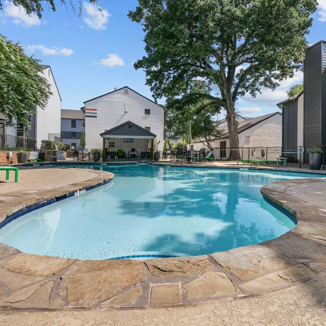 A residential swimming pool surrounded by green trees and buildings, with lounge chairs nearby.