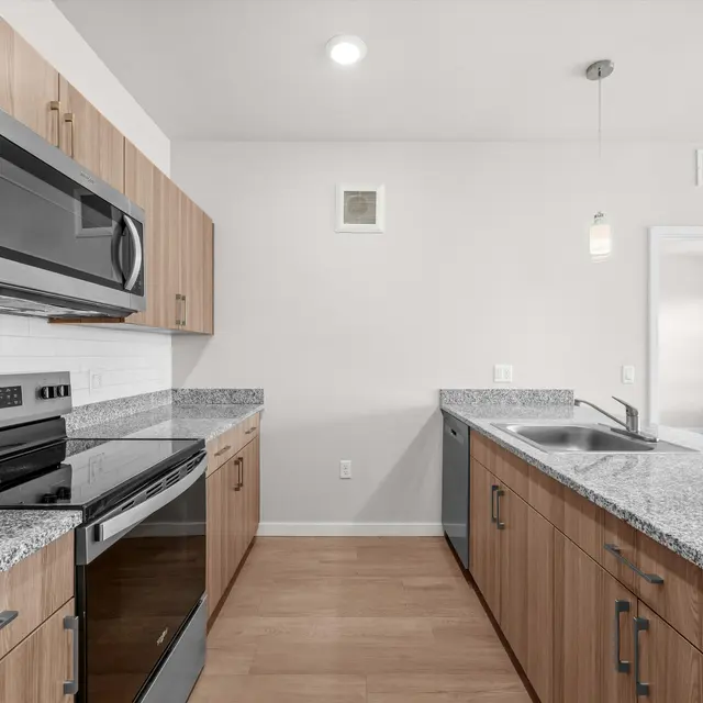 A modern kitchen featuring wooden cabinets, stainless steel appliances, and granite countertops. The kitchen is well-lit with pendant lights above the countertop and a large window allowing natural light in.