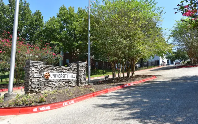 A stone entrance sign for University Hill, surrounded by trees and a paved road in a sunny landscape.