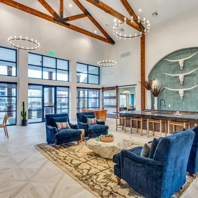 A spacious modern lobby featuring high wooden beams, blue upholstered chairs, a patterned rug, and a stone counter area with decorative elements.
