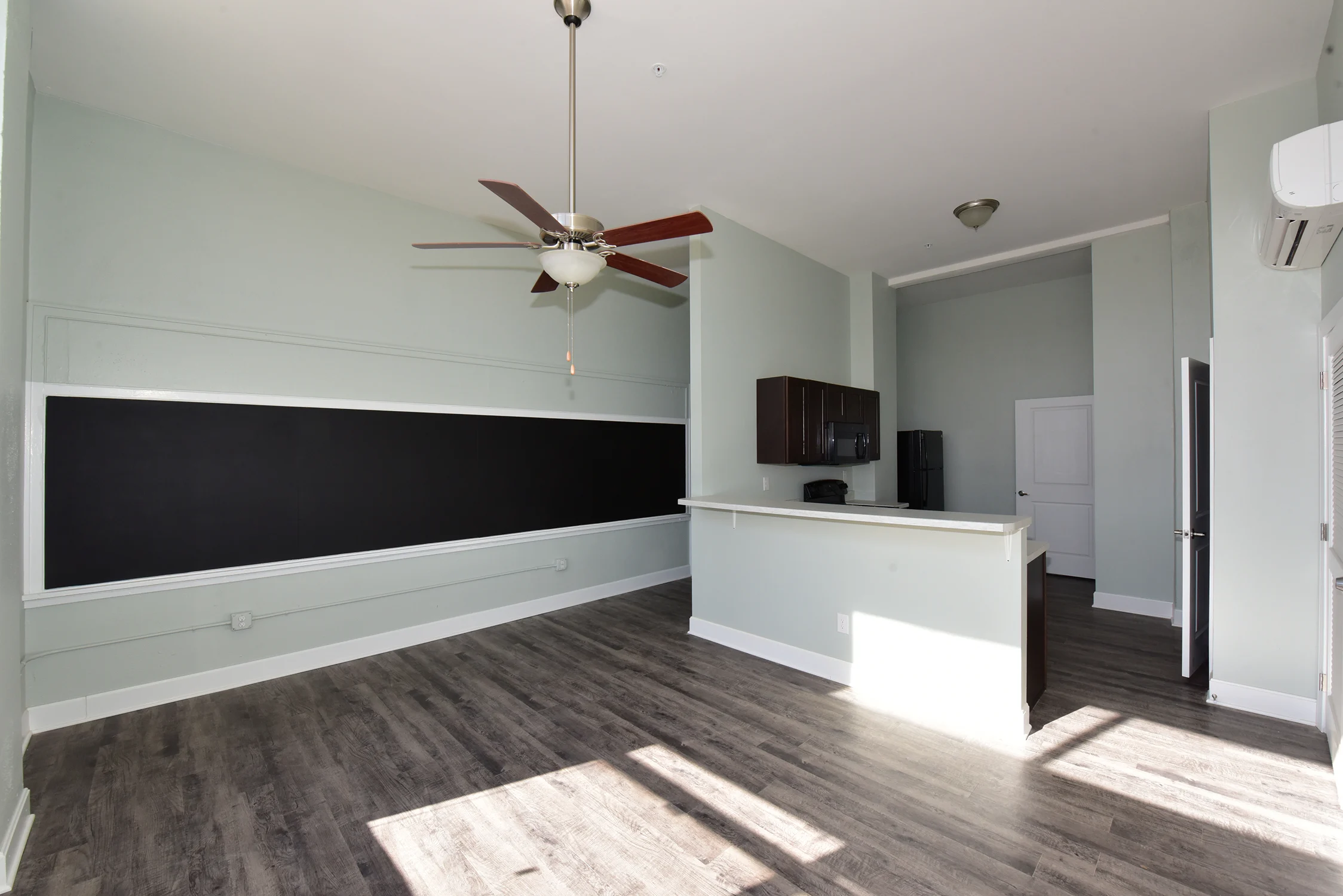 Modern Apartment Interior Spacious modern apartment interior with a ceiling fan, light green walls, and wooden flooring. A large chalkboard wall is visible, along with a kitchen area showcasing dark cabinets and appliances.