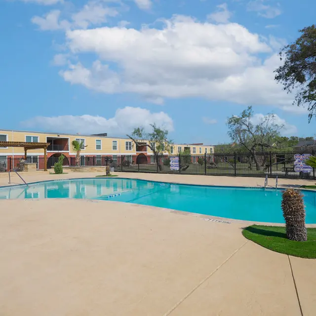 A view of a clean, blue swimming pool surrounded by a landscaped area and apartment buildings in the background under a clear sky.