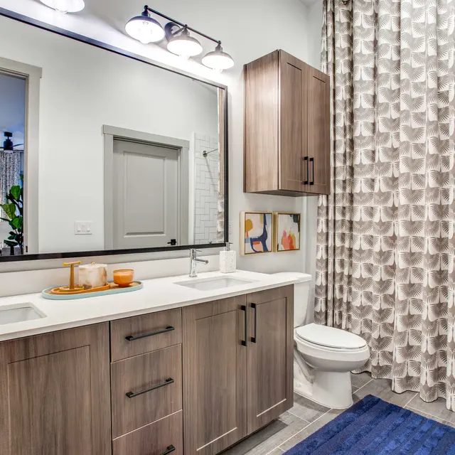 A modern bathroom featuring a double sink vanity with wooden cabinets, a large mirror, and stylish fixtures. The bathroom has a shower and bathtub, with a patterned shower curtain and a blue bath mat.