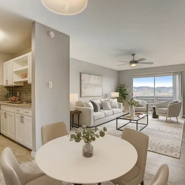 A modern living room and kitchen setup in an apartment. The kitchen features white cabinetry, a small seating area with a table and chairs, and an open view into a well-furnished living space. The living area has a sofa, armchairs, a coffee table, and large windows providing natural light and a scenic view.