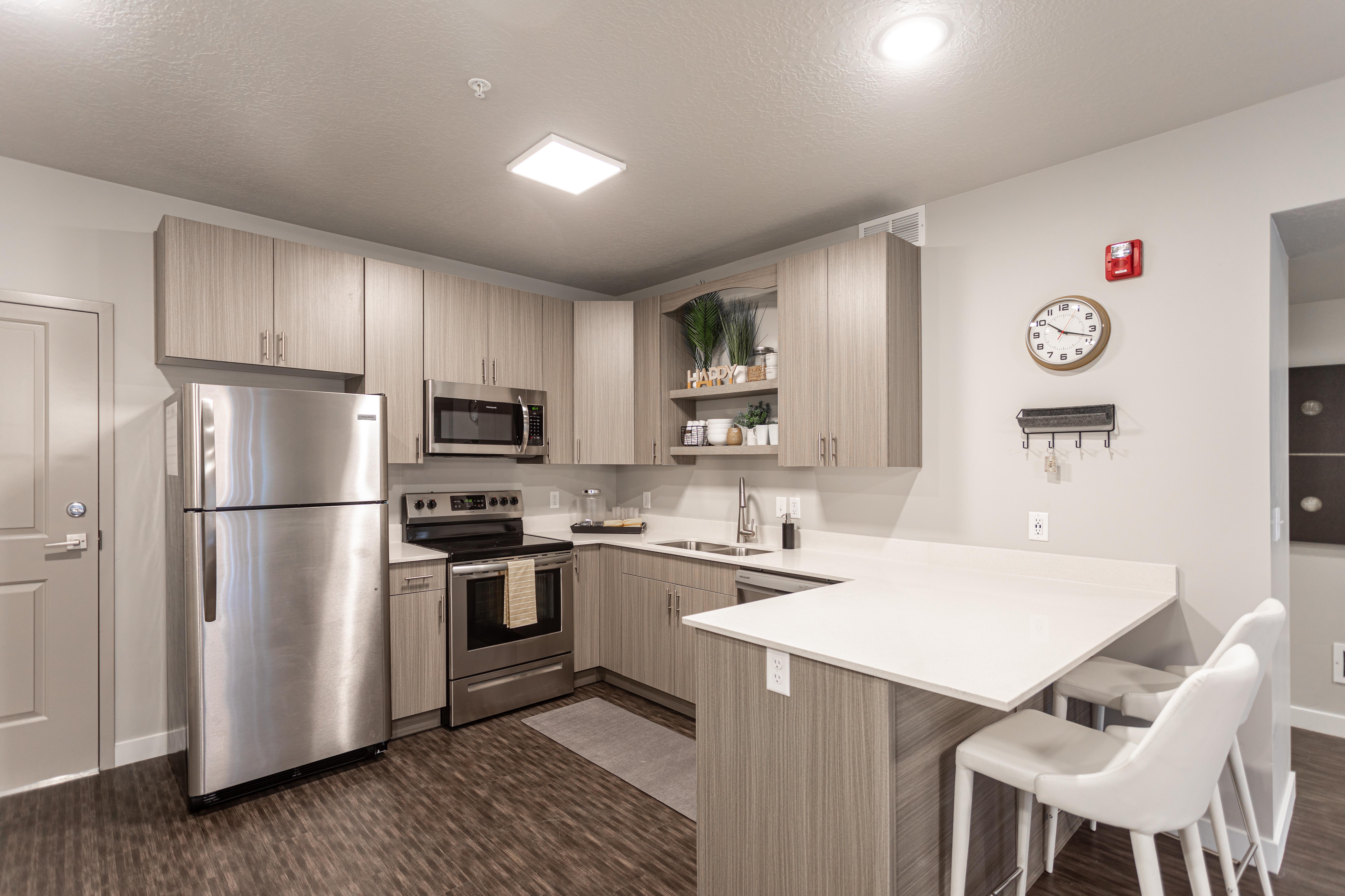 A modern kitchen featuring stainless steel appliances, light wooden cabinetry, and an island with seating. The decor is minimalist with plants and kitchen essentials displayed on shelving.