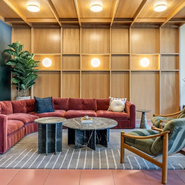 A cozy and modern living room with a large red sectional sofa, a circular dark marble coffee table, and two wooden accent chairs. The wall features a wooden panel with round lights, and a plant adds a touch of greenery.