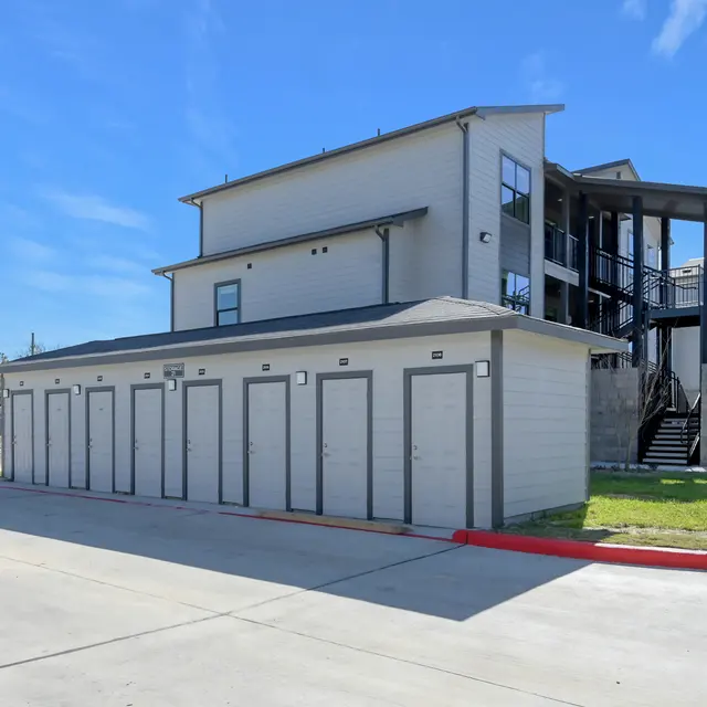 A modern storage facility with multiple storage units in front and a multi-story residential building behind it. The sky is blue with a few clouds.