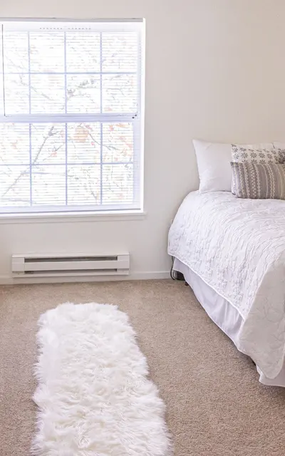 A cozy bedroom featuring a bed with light-colored bedding, a set of decorative pillows, a soft white rug on the beige carpet, and a window with blinds allowing natural light to fill the room.