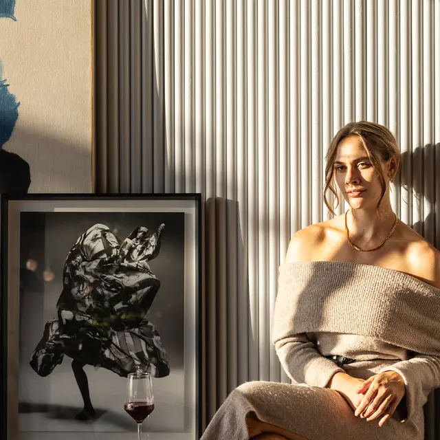 A woman in a cozy, off-shoulder sweater sitting beside a framed artwork and a glass of wine, with soft shadows and light creating a warm atmosphere.
