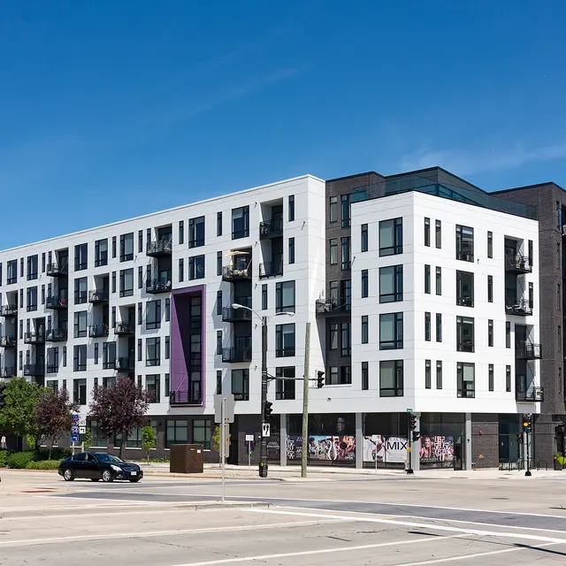 A modern, multi-story apartment building with a combination of white and dark exterior, featuring balconies and large windows. The architecture is contemporary with a mix of materials. The building is situated on a city street with clear blue skies in the background.