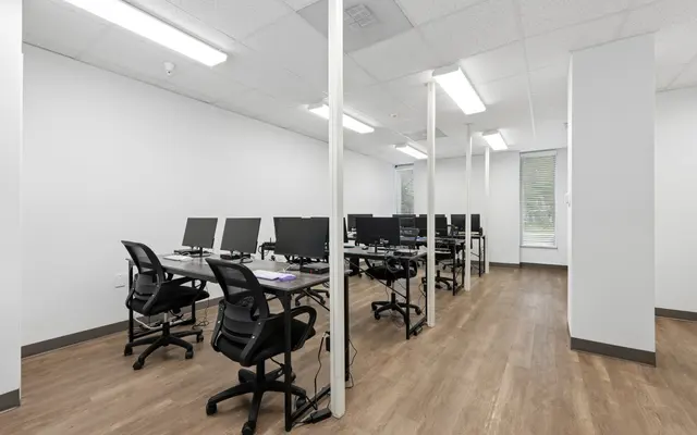 A spacious, modern office with multiple desks and computer monitors arranged in rows. The floor is wooden and the walls are white, creating a clean and minimalistic atmosphere.