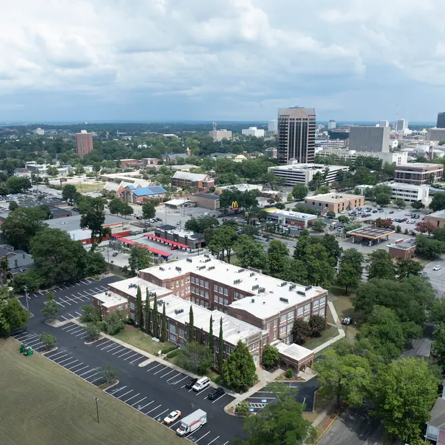 An aerial view of a city with a mix of low-rise buildings, parking lots, and green trees, under a partly cloudy sky.