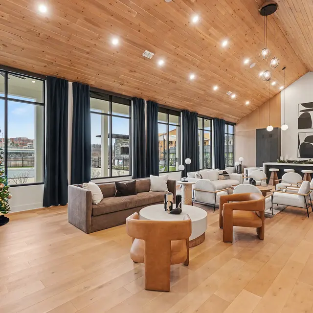 A spacious modern living room featuring large windows, wooden ceiling, and a Christmas tree. The room includes a couch, chairs, and decorative lighting.